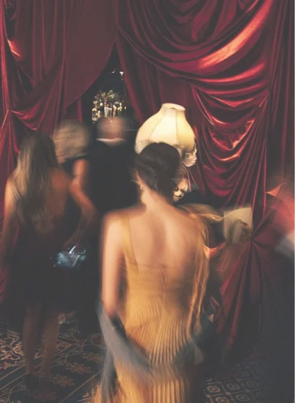 A motion-blurred, candid photograph capturing the dynamic energy of a formal gathering or party. Guests in elegant evening wear, including a woman in the foreground wearing a golden backless dress, move past a dramatic backdrop of rich, draped crimson velvet curtains. A vintage fringed lamp glows warmly in the center of the scene.