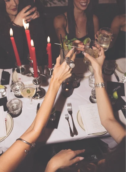 Friends raising glasses in a celebratory toast around an elegantly set dining table with red candles, cocktails, and dinnerware.