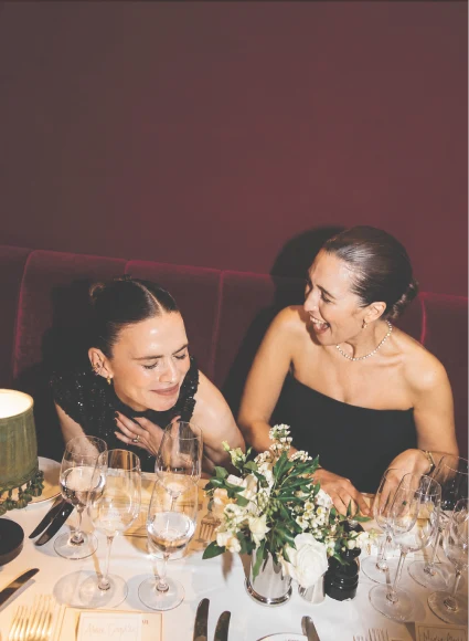 Two women smiling and enjoying a meal together at an elegantly decorated dinner table with floral centerpieces and wine glasses.