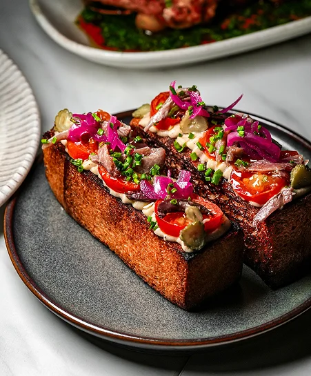 High-angle professional food photography of a ceramic platter featuring four unique gourmet artisan toasts. From left to right: pickled red onions with savory toppings, sliced fresh figs with goat cheese and walnuts, smoked salmon with red onion rings and capers, and smashed avocado topped with a poached egg and microgreens.