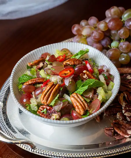 White ceramic bowl filled with halved red grapes, pecans, sliced chili peppers, and herbs served on a silver platter.