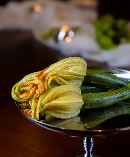 Fresh squash blossoms with attached small baby zucchini resting on a polished metal pedestal dish