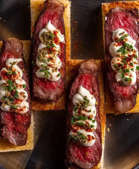 Seared beef steak slices on toasted crostini topped with whipped cream, paprika, and chopped chives.