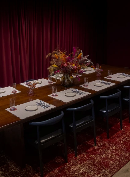 Long wooden dining table set for a group with placemats, plates, cutlery, drinking glasses, dark blue chairs, red drapes, and a central dried flower arrangement.